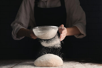 Woman sprinkling flour over dough at wooden table on dark background, closeup