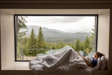 Woman lying on window sill covered with blanket, enjoying great view on mountains and pine forest...