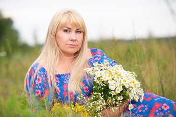 Naklejka na ściany i meble A beautiful plus size girl with white hair in a summer dress poses on the street with daisies. Chubby girl in a meadow with daisies close-up.