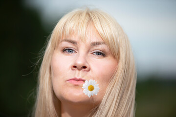 A middle-aged woman with white hair holds a chamomile flower in her mouth.