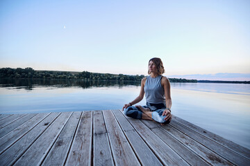 A yogi woman sits on a dock in a lotus pose and meditates.