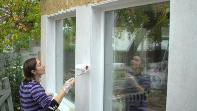 Woman paints the wall between the windows of her own house outdoors