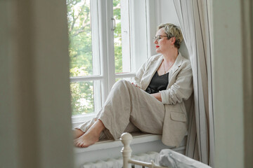 A middle-aged woman in a beige suit and black tank top sits mysteriously and looks out the window on the windowsill. Green trees outside