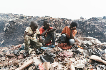 Group of African boys sitting among garbage and plastic waste in an illegal landfill; health risk...