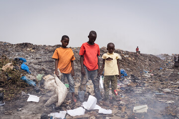 Group of poor African boys looking for recyclable items standing together amid burning garbage in...