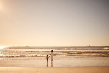 Fototapeta na wymiar Girl and mother walking in the water at the beach during sunset on summer family vacation. Young daughter and her mom on sand together by the ocean during the dusk on travel holiday with mockup space