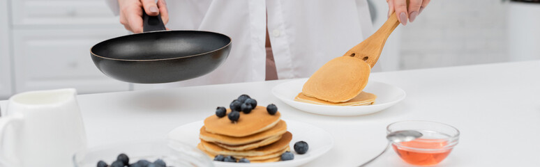 Cropped view of woman in shirt putting pancake on plate near fresh blueberries in kitchen, banner.