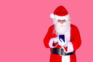 Fototapeta na wymiar Funny old bearded Santa Claus in a suit holding a mobile phone with a mobile app on a smartphone, shocked by a Christmas advertisement isolated on a pink background.