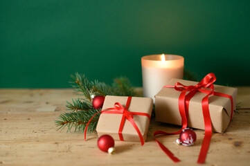 Close up of fancy candle with Christmas gift boxes with red ribbons, natural pine tree pine branches on brown wooden table in front of green background