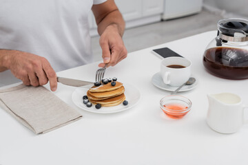 Cropped view of man cutting pancakes near coffee and smartphone in kitchen.