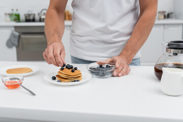 Cropped view of man putting blueberries on pancakes near coffee pot in kitchen.
