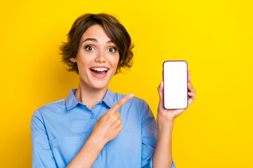 Portrait of impressed excited girl with brown hair dressed blue shirt indicating at phone app advert isolated on yellow color background