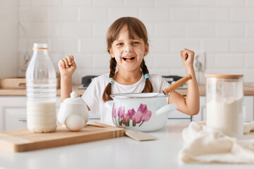 Horizontal shot of excited amazed child in a kitchen making a dough, sitting at table surrounded with products and tableware, wants to bake a pie, clenched fists, celebrating her success in baking.