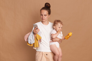 Indoor shot of serious young adult woman with bun hairstyle wearing white t shirt standing with her toddler daughter and using mobile phone with concentrated expression, isolated over brown background