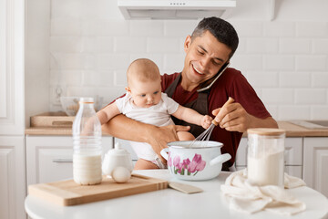Obraz na płótnie Canvas Indoor shot of brunette man in maroon t shirt and apron sitting at table with infant daughter in kitchen, talking phone with positive expressing and mixing dough.