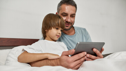 Father and little son watch tablet in bed at home