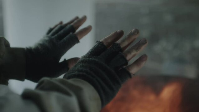 Close up shot of hands of unrecognizable homeless man in fingerless mittens warming near fire barrel