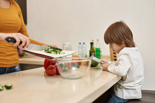 Boy Use Tablet While Mom Pour Cut Cucumber In Bowl