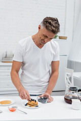 Man putting fresh blueberries on pancakes near coffee and honey in kitchen.