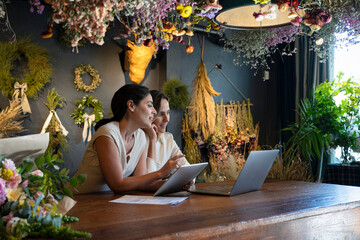 Wide view of two diverse female home decoration shop owners working with laptop computer and digital tablet