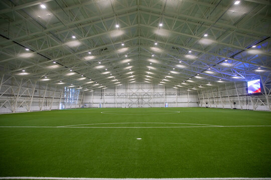 A New Empty European Sports Hall For Playing Football At Any Time Of The Year. The Football Field Is Covered With Artificial Grass, Lighting Spotlights Are Installed On The Walls And Ceiling.