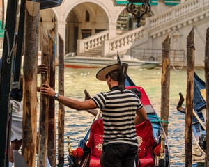 The gondolier helps tourists enter the gondola near the bank of the canal of the city of Venice on a sunny morning, the Venetian gondolier extended a helping hand to the passengers of the boat