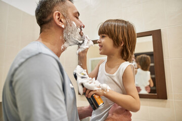 Boy smear shaving foam on father beard at bathroom