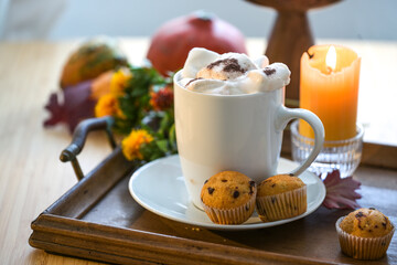 White mug with chocolate hot drink and melting marshmallows, muffins, candle and autumn decoration on a table, copy space, selected focus