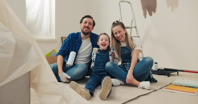 Happy family in new apartment. Sitting on floor is a handsome middle-aged man beautiful wife and their very cute daughter. A man hugs daughter, who is wearing denim overalls and a checkered shirt.