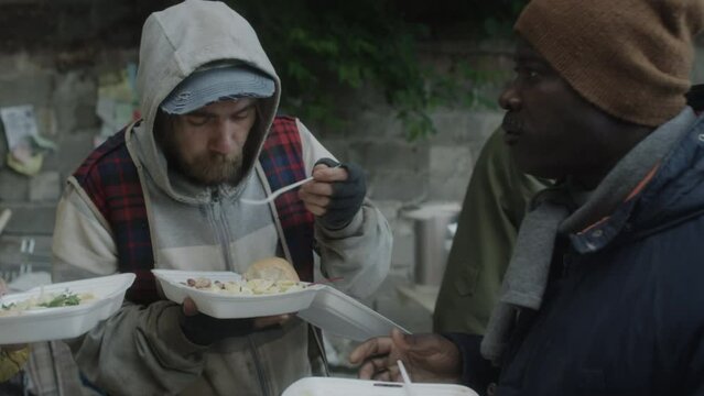 Group of multiethnic homeless men talking while eating free meal served by volunteers on outdoor soup kitchen
