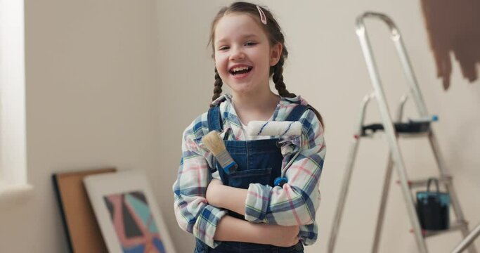 Cute girl with brown hair and gray eyes wearing denim overalls and checkered shirt holding paint roller. Child has paintbrush in pocket. She is looking forward to painting wall in living room herself.