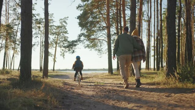 View from the back parents dad and mom walk together and the daughter in front rides a bicycle in a helmet. A sei walk in the woods to the lake. Happy family with a bicycle