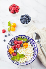 Cottage cheese in bowl garnished with raspberries, blueberries, physalis and mint leaves on white background