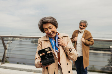 cheerful senior woman holding vintage camera near blurred husband on background.