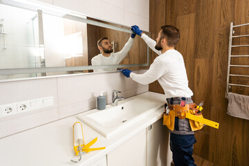 The worker installs the mirror in the bathroom. - obrazy, fototapety, plakaty