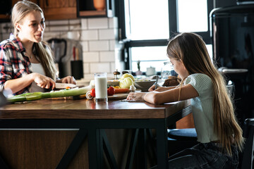 Mother prepares ingredients for making the meal while her daughter stands by the kitchen table and drinks milk.	