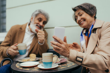 happy senior woman holding smartphone while having brunch with husband on terrace of cafe.