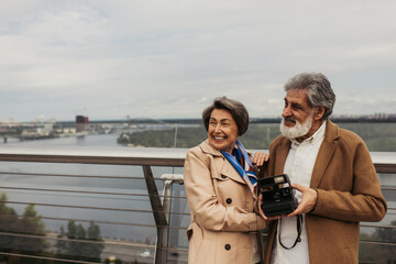 happy senior woman in trench coat hugging husband with vintage camera standing outside.