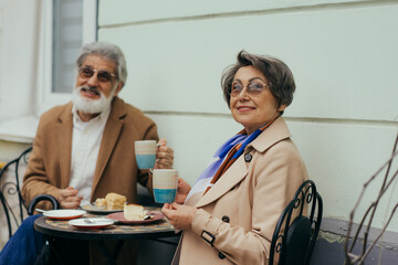 happy senior woman in eyeglasses holding cup while having brunch with bearded husband on terrace.