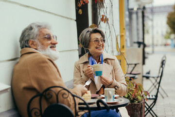 cheerful senior woman in eyeglasses and trench coat holding cup during brunch with husband on terrace of cafe.