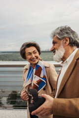 happy senior man holding vintage camera near curious wife smiling on bridge.