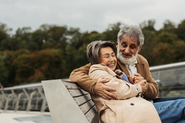 Fototapeta premium happy senior man hugging joyful wife while sitting on bench outdoors.