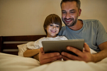 Smiling father and son watch digital tablet in bed