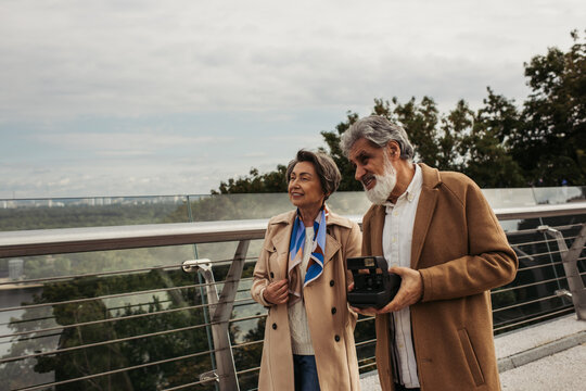 happy senior man holding vintage camera near cheerful wife on bridge.