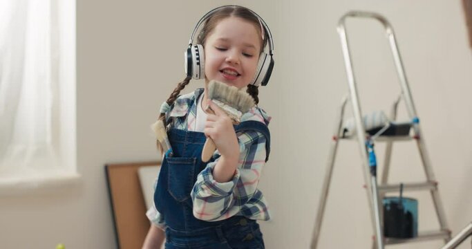 Girl listens to music in headphones and sings paint roller. Kid is wearing denim overalls and checkered shirt. In background there is ladder and a dustpan, as the apartment is under renovation.
