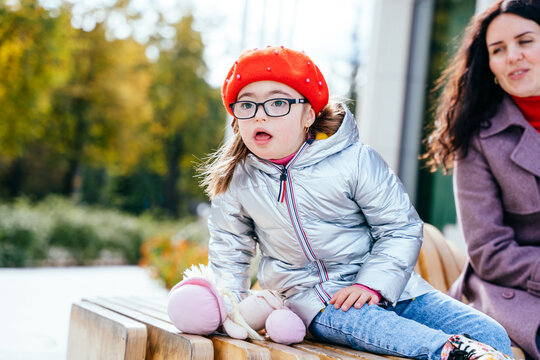 Happy family moments in the city. Cute curious little daughter in eyeglasses wearing red beret and mother are sitting at bench outdoor in the city street.