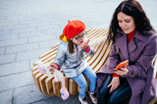 Brunette caucasian positive woman and daughter using smartphone sitting on the bench at city street. Mother calms the girl showing cartoon on the phone.