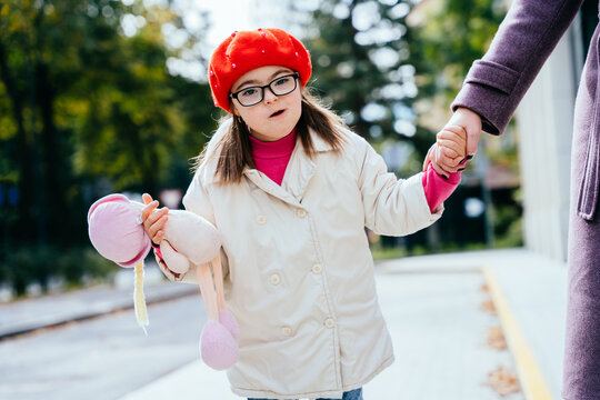 Cute girl in eyeglasses wearing coat red beret with special needs holding mother's hand on a walk outdoor.