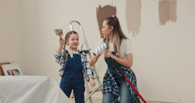 A blonde woman and a cute 8-year-old girl are dancing in the middle of a renovated room. Instead of microphones, they have paint rollers and brushes in their hands.