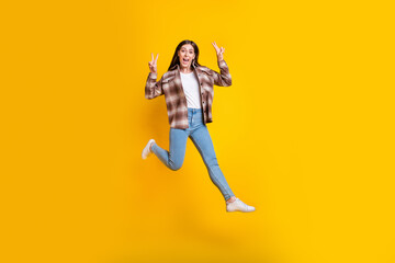 Fototapeta na wymiar Full length photo of pretty shiny girl dressed plaid shirt jumping high showing v-signs isolated yellow color background